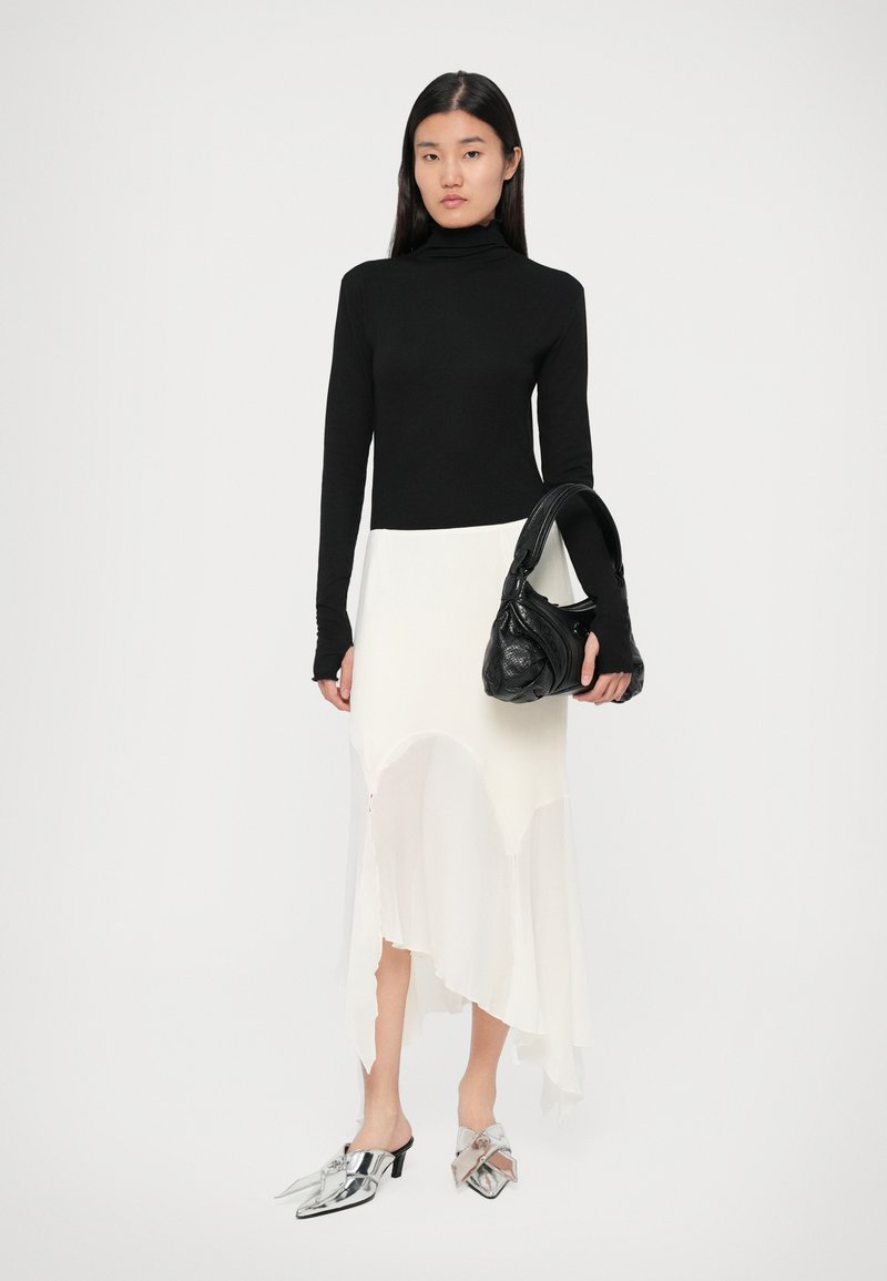 Schwarzer Rollkragenpullover, weiße asymmetrische Schürze, silberne Absatzmules und eine strukturierte schwarze Tasche mit einem gebogenen Griff.