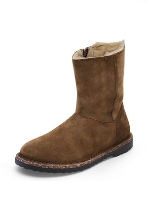 Stivaletto in suede marrone con morbida foderatura in pelliccia, texture liscia, punta arrotondata e suola in gomma texturizzata per una migliore aderenza. Chiusura con zip sul lato.