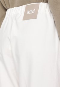 Hvite stoffbukser med elastisk midjebånd, med en beige etikett med "MM"-logo bak. Glatt tekstur, uformell design.