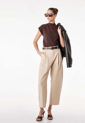 Frau trägt übergroße beige Hose, dunkelbraunes ärmelloses Top, große Sonnenbrille, schwarze Lederjacke über der Schulter tragend, stehend.