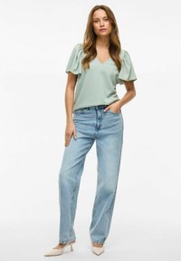 Svijetlozelena bluzica s V izrezom i puf rukavima, uparena s visoko strukiranim, širokim svijetloplavim trapericama i bež cipelama s oštrim nosom.