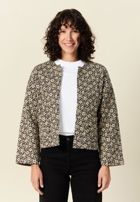 Czarna i kremowa kurtka w kwiatowy wzór, krótka konstrukcja z długimi rękawami i okrągłym dekoltem, noszona na białej koszuli i czarnych spodniach.
