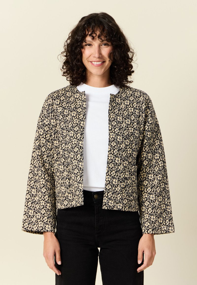 Czarna i kremowa kurtka w kwiatowy wzór, krótka konstrukcja z długimi rękawami i okrągłym dekoltem, noszona na białej koszuli i czarnych spodniach.