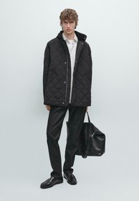 Zwarte gewatteerde jas met een kraag, gecombineerd met een witte shirt, donkere spijkerbroek en een zwarte leren tas. De look wordt compleet gemaakt met zwarte schoenen.