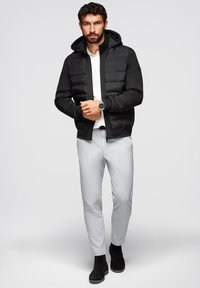 Giubbotto puffy nero con cappuccio, in materiale poliestere, con polsini e orlo a coste, indossato sopra una camicia bianca, abbinato a pantaloni grigi chiari e stivali neri.