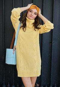 Abito giallo a maglia con colletto alto a costine, caratterizzato da un design a treccia. Abbinato a una borsa azzurro chiaro e a un cerchietto arancione.