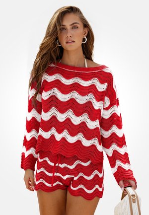 Femme portant un pull en maille rayée ondulée rouge et blanche et un short, tenant un sac à main en paille tressée, avec des boucles d'oreilles créoles en perles.