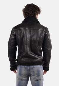 Giubbotto in pelle nera con finitura testurizzata, dotato di colletto in shearling e cinturini laterali regolabili. Indossato sopra jeans blu sbiaditi.