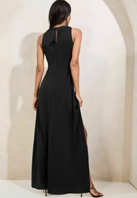 Rochie maxi neagră cu un design fără mâneci, fante laterale și un decolteu în formă de cheie pe spate. Textură de material neted, croială până la podea și accesorii minime.