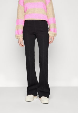 Personne portant un pantalon noir évasé, un pull rayé beige et rose, et des baskets compensées blanches, debout sur un fond uni.