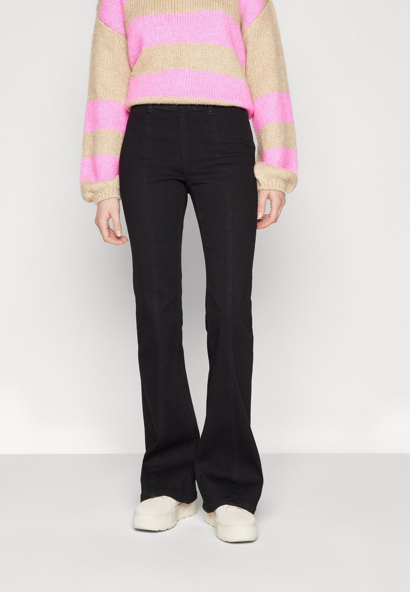 Personne portant un pantalon noir évasé, un pull rayé beige et rose, et des baskets compensées blanches, debout sur un fond uni.