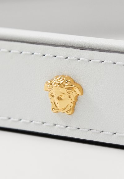 Balta oda su auksiniu Medūzos emblema, išsamiai siuvinėta ir glotnaus paviršiaus. Akcentai pabrėžti kontrastinga tamsia riba.