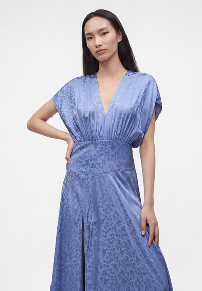 Femme aux longs cheveux foncés portant une robe en satin bleu clair à motif floral, décolleté profond en V, large ceinture et fente avant, posant avec une main sur la hanche.