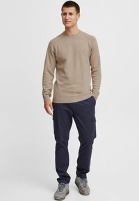 Beige strikket sweater med rund hals, parret med mørkeblå cargo-bukser med sidelommer. Skoene er grå med accenter.