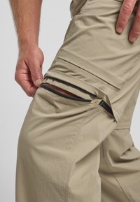 Beige Outdoor-Hosen aus strukturiertem Stoff mit einem seitlichen Reißverschlussfach. Das Fach ist mit schwarzem Zubehör ausgestattet und hat ein glattes, funktionales Design.