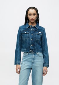 Lühendatud teksajakk tumedas sinises, klassikalise krae, ees olevate nuppude ja kahe rinnataskuga. Kandmiseks valitud helesiniste kõrge vöökohaga teksadega.