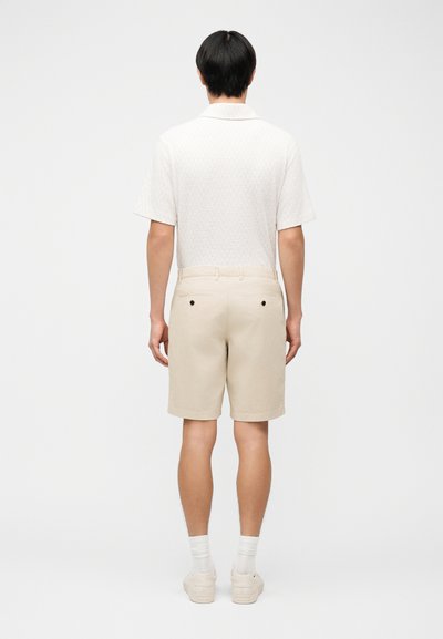 Άνδρας που φοράει μπεζ σορτς, λευκό κοντομάνικο πουκάμισο, λευκές κάλτσες και μπεζ παπούτσια, στέκεται με την πλάτη προς τα εμπρός σε απλό φόντο.