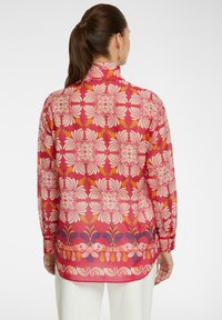 Camicia a maniche lunghe di un vivace colore rosa con motivi floreali bianchi e arancioni, con colletto e orlo curvo, realizzata in tessuto leggero.
