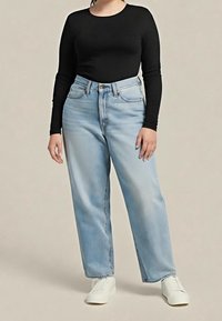 Femme portant un haut noir à manches longues, un jean taille haute bleu clair et des chaussures blanches, debout devant un fond uni.