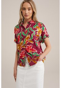 Cămașă cu mânecă scurtă, fabricată dintr-un material colorat, având modele florale mari în roșu, galben, verde și purpuriu pe un fundal adânc de vișiniu.