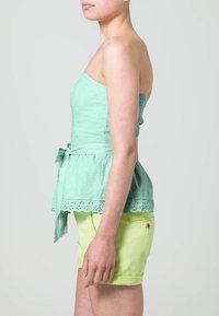 Top senza spalline verde menta con design a occhio di pernice, vita peplum e fascia annodata, abbinato a pantaloni corti arrotolati giallo chiaro.