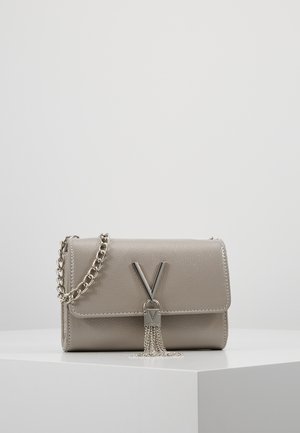 Mała szara skórzana torebka z srebrnym łańcuszkiem, metalowym zapięciem w kształcie litery V oraz zwisającymi srebrnymi frędzlami na białej powierzchni.