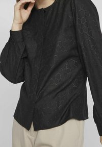 Donna che indossa una blusa nera a maniche lunghe con motivi floreali ad occhiello e pantaloni beige, con una mano vicina al viso.