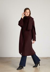 Bordeaux wollen jas met een hoge kraag, voorzien van drie grote zwarte knopen, zijsplitten en lange mouwen, gecombineerd met flare jeans en enkellaarsjes.