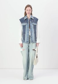 Denim vest with a blue patterned front, white shirt underneath, and light blue flared pants featuring cutouts. Model holding a small bag.