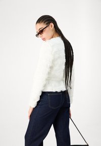 Eine Frau mit langen, geflochtenen Haaren, die dunkle Sonnenbrillen trägt, eine strukturierte weiße Jacke, dunkelblaue Jeans und eine schwarze Handtasche hält.