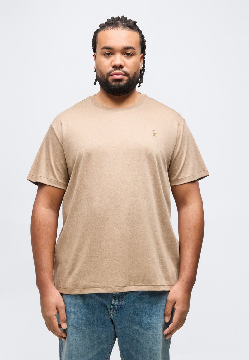 Άνδρας με πλεξούδες στα μαλλιά και γενειάδα, φορώντας μπεζ κοντομάνικο μπλουζάκι και μπλε τζιν, στέκεται μπροστά σε απλό ανοιχτό φόντο.