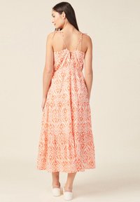 Maximekkopuku oranssi-valkoisessa kukkakuosissa, jossa on ohuet säädettävät hihnat, avainreikäselkä ja kerroksellinen röyhelöhelma. Litteät valkoiset kengät.