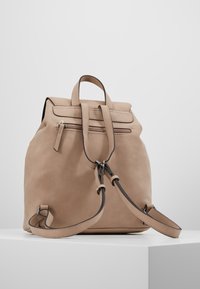 Beige mockaskinnsryggsäck med ett övre handtag, justerbara axelremmar och en dragkedjefrontficka. Har sömnadsdetaljer och en mjuk textur.