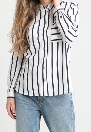 Vrouw draagt een wit overhemd met verticale zwarte strepen en lichtblauwe jeans, haar hand rustend vlakbij haar gezicht.