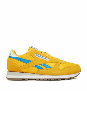Gelber Reebok-Sneaker mit blauen Seitenstreifen, weißer Sohle und Logo an der Seite und Ferse, von der Außenseite auf weißem Hintergrund gezeigt.
