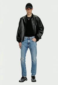 Svart läderbomberjacka över en svart t-shirt, ljusblå slitna jeans och svarta skor. Modellen bär en svart keps och ett silverhalsband.