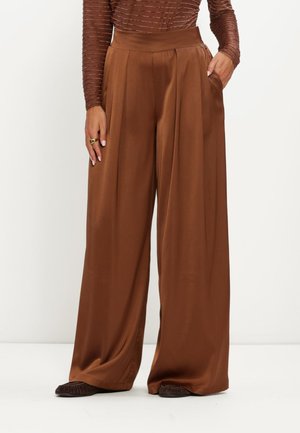 Vrouw die hoge taille wijde bruine satijnen broek met plooien draagt, gecombineerd met een bruin langarmig shirt en donkere schoenen, staand met één hand in de zak.