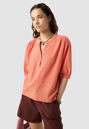 Blouse corail à manches courtes avec une patte de boutonnage semi-longue et des épaules froncées, assortie à un short marron. Texture lisse, coupe décontractée.