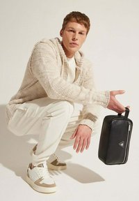 Borsa in pelle nera di forma rettangolare, con cerniera argentata e manico, tenuta da una persona in un maglione crema e pantaloni bianchi, accucciata.