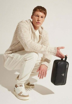 Borsa in pelle nera di forma rettangolare, con cerniera argentata e manico, tenuta da una persona in un maglione crema e pantaloni bianchi, accucciata.