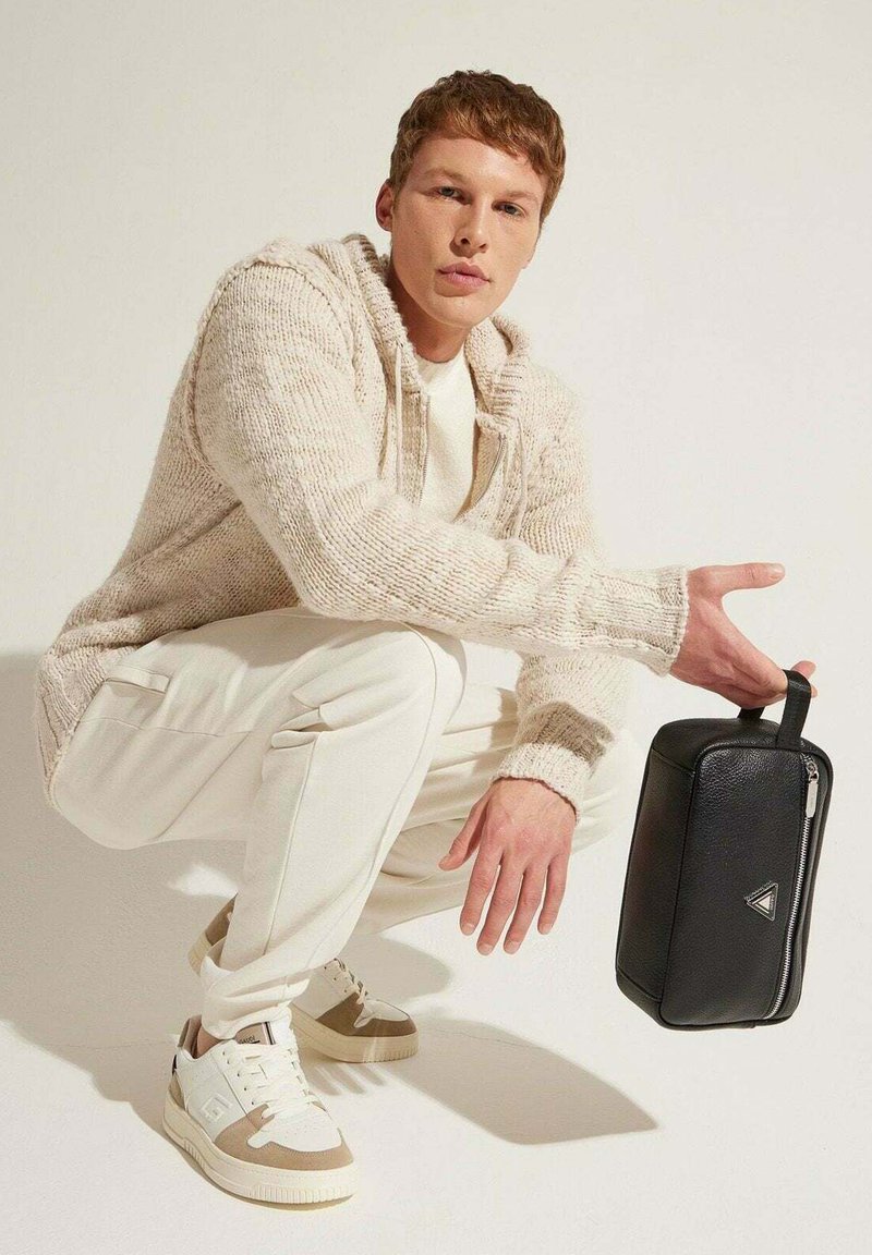 Borsa in pelle nera di forma rettangolare, con cerniera argentata e manico, tenuta da una persona in un maglione crema e pantaloni bianchi, accucciata.