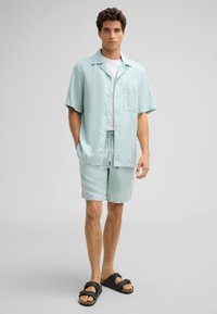 Hellblaues kurzärmliges Hemd mit Knopfverschluss, kombiniert mit passenden Shorts und schwarzen Sandalen. Glattes Material mit lässiger Passform.