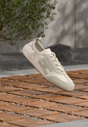 Adidași casual albi și bej cu șireturi, vârf din piele întoarsă și un emblemă mare "H", poziționați deasupra unei suprafețe exterioare cu plăci de lemn, cu fundal de verdeață.