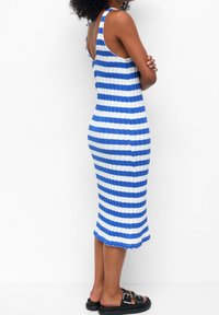 Abito lavorato a maglia con strisce orizzontali blu e bianche, design aderente, lunghezza fino al polpaccio, spalline larghe e texture a coste. Sandali neri aperti.