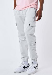 Uomo che indossa pantaloni cargo jogger grigi chiari, sneakers bianche Nike Air Force 1 e una maglietta rosa chiaro, in piedi con le mani nelle tasche.