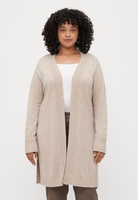 Svetlo béžový cardigan, pletený materiál, otvorený design, rebrované manžety a spodný lem, siaha po strednú časť stehien, nosený cez biele tričko a hnedé nohavice.