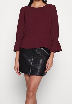 Femme portant un chemisier bordeaux à manches longues avec des poignets évasés et une mini-jupe en cuir noire avec des fermetures éclair argentées en diagonale.