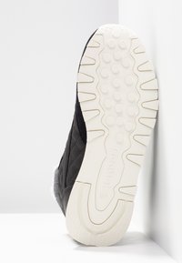 Svart vadderad sneakersula med vit texturerad sula, som visar cirkulära grepp och Reebok-logotyp, uppställd mot vit vägg.