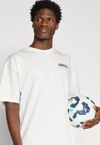 T-shirt de algodão branca com mangas curtas, apresentando "JUVENTUS" e "MILLESST" impressas em preto e rosa. Segurando uma bola de futebol branca com acentos azuis.