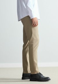 Beige cotton trousers with a straight-leg cut, paired with black leather shoes, shot from the side against a plain background.